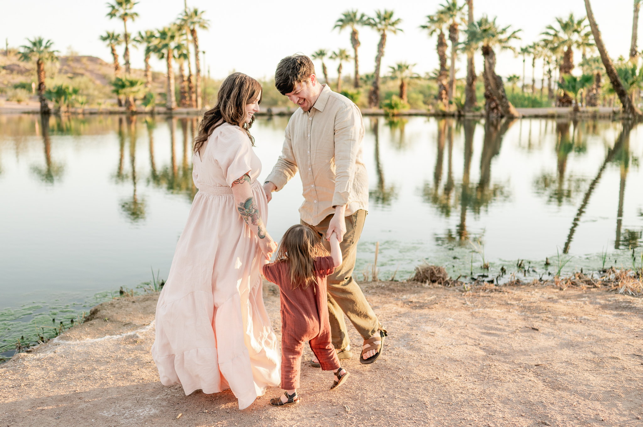 pregnant mama and her husband and daughter dance at the water's edge at papago park with palm trees and the lake behind them