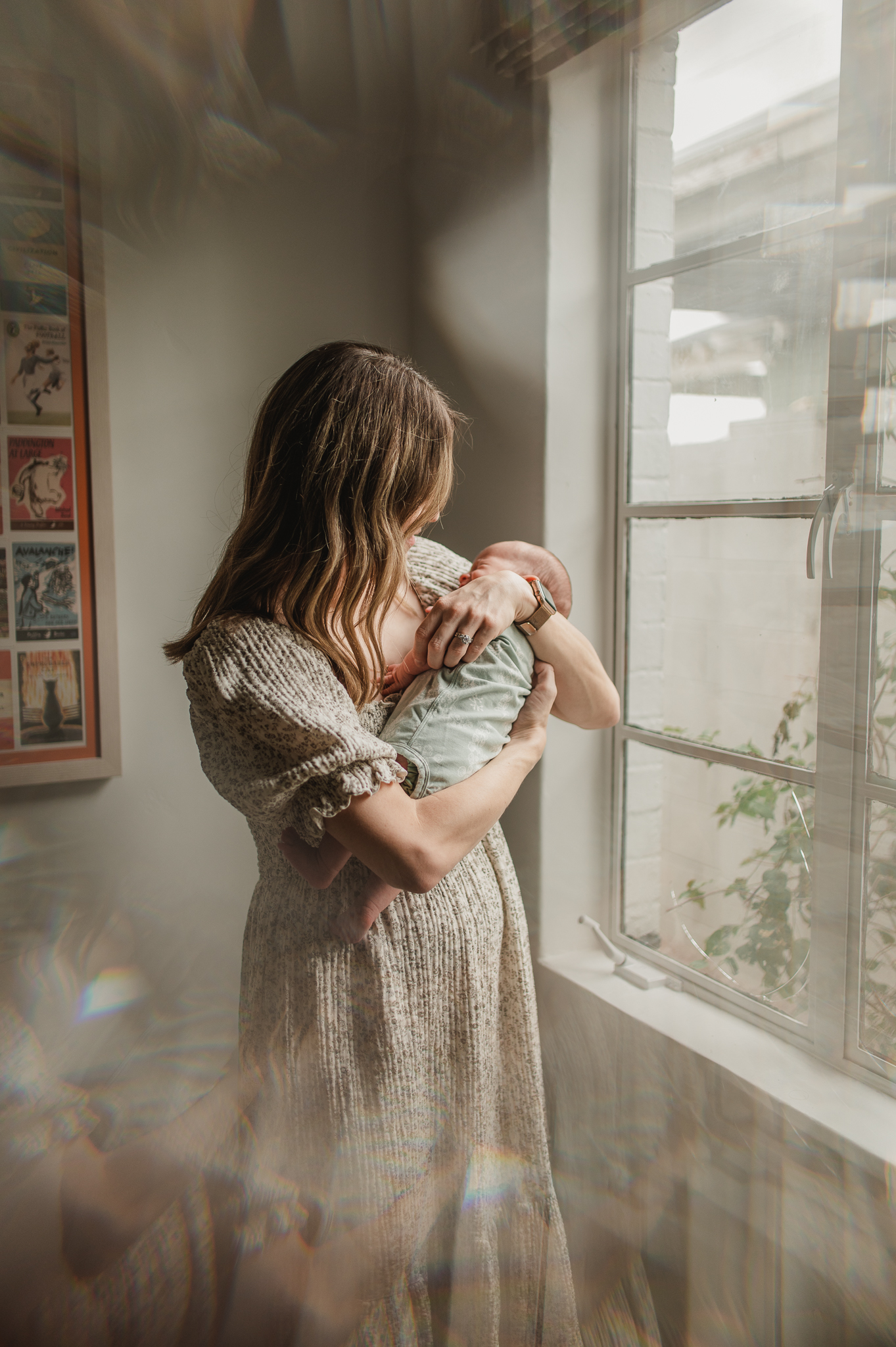 mother in cream floral dress holding her newborn baby girl next to her nursery window
