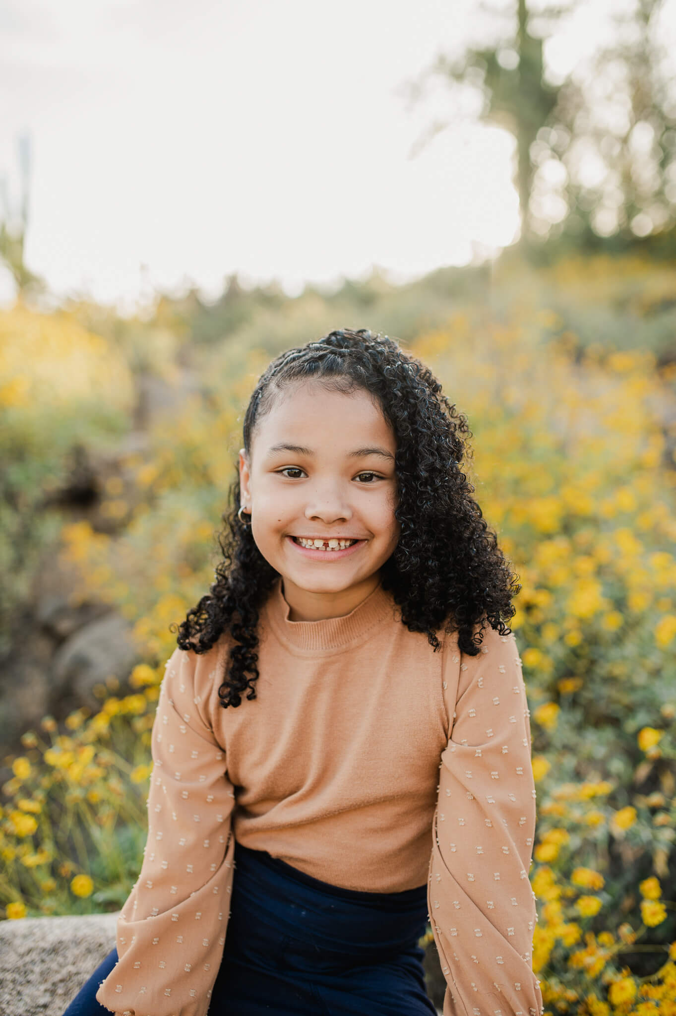 young girl wearing a camel sweater smiling at the camera sitting on a boulder amongst a field of Phoenix wildflowers