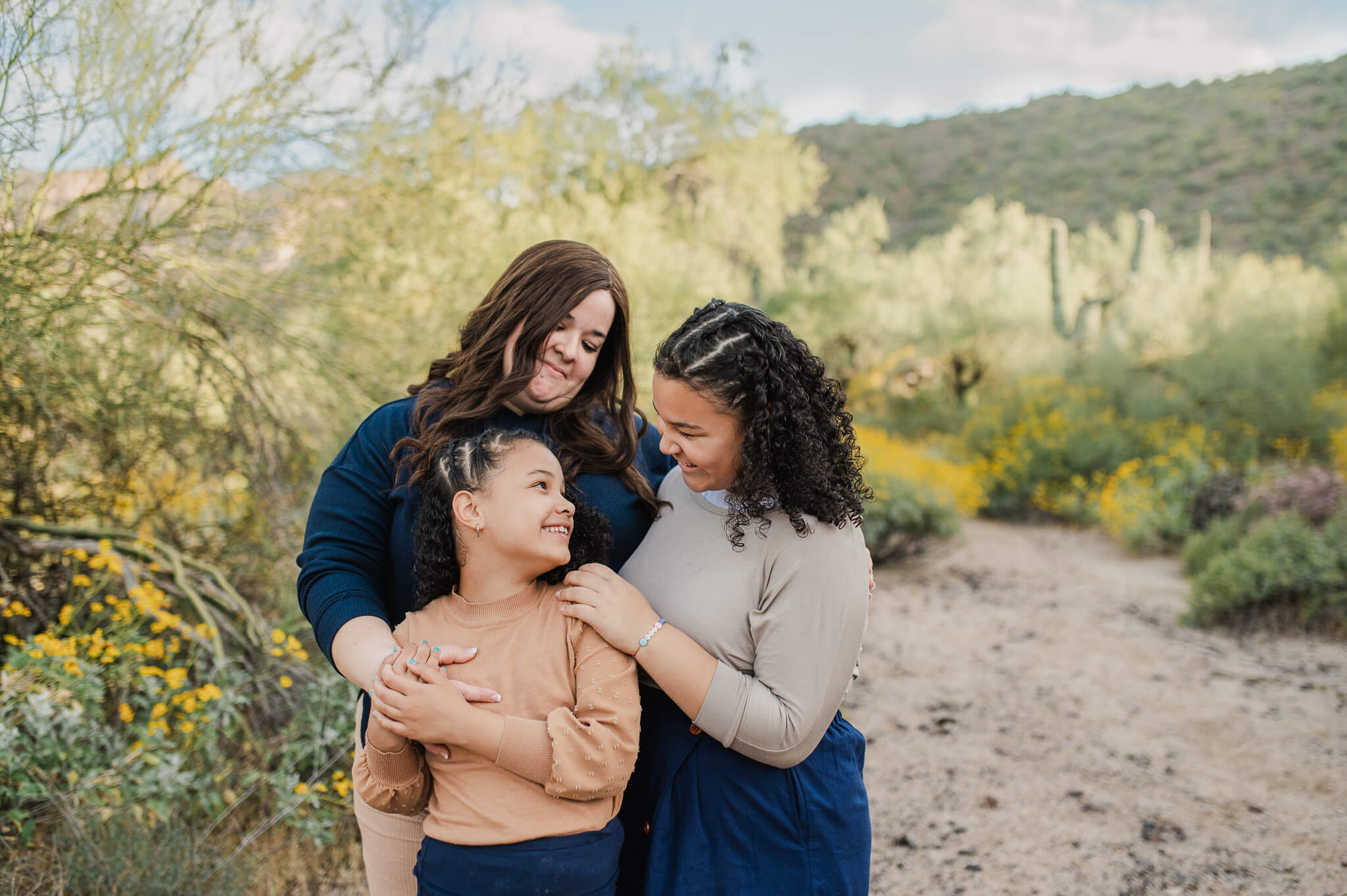 Mom holding her older and younger daughter in her arms while they stand in front of a path surrounded by Phoenix wildflowers