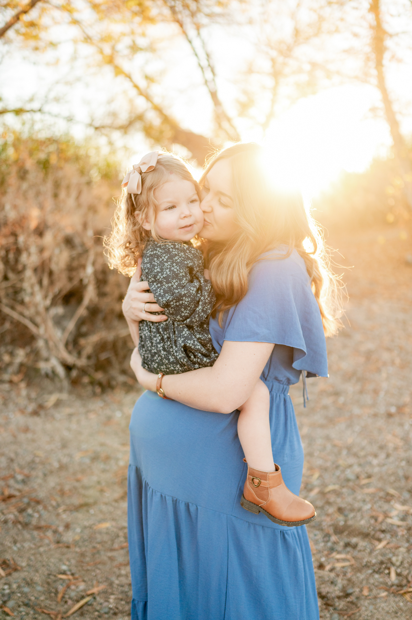 pregnant mom in a blue dress kissing her 3 year old daughter who's sitting on her belly, with a sun flare shining behind them
