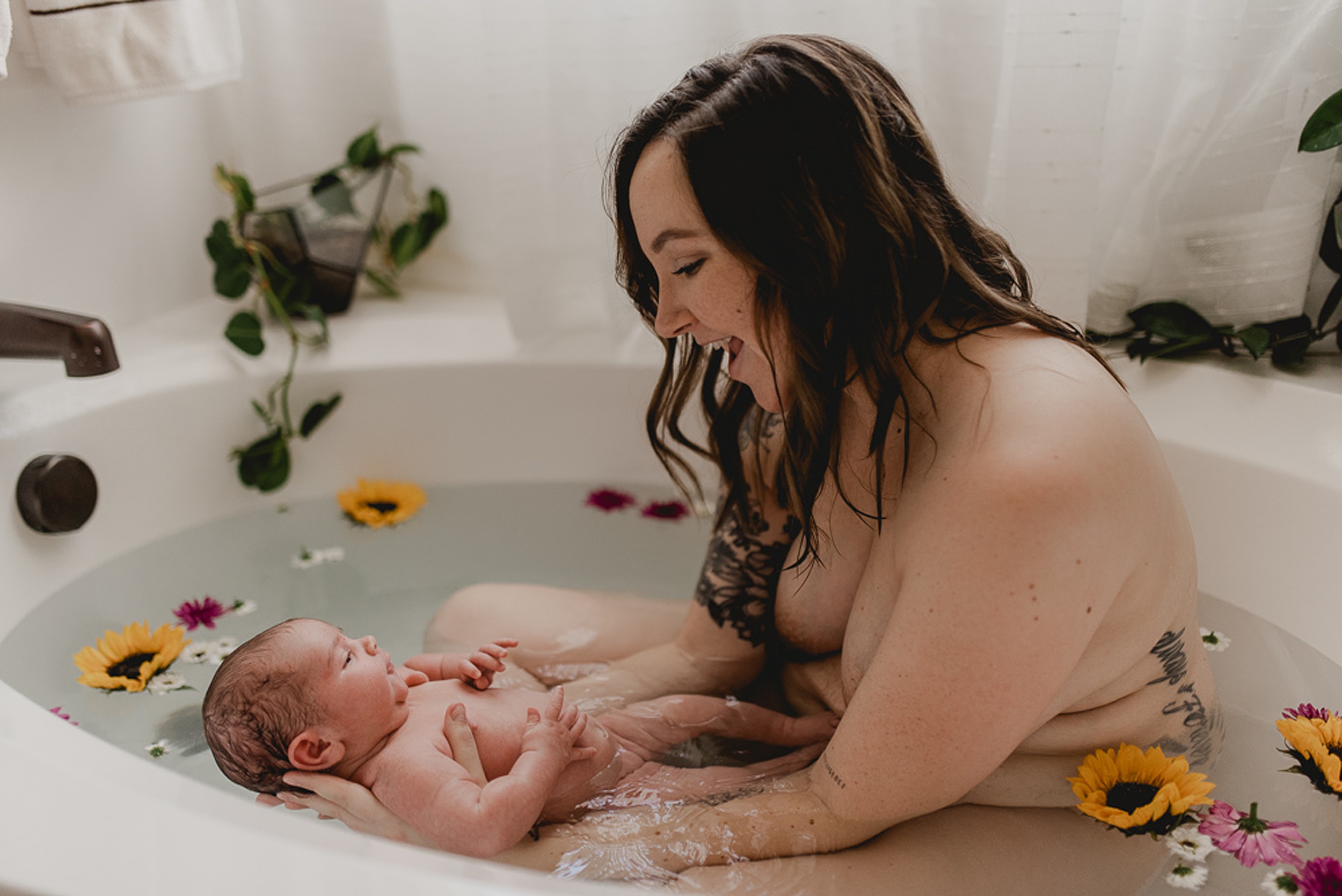 A new mom laughs and plays with her newborn baby while sitting in a bathtub surrounded by flowers