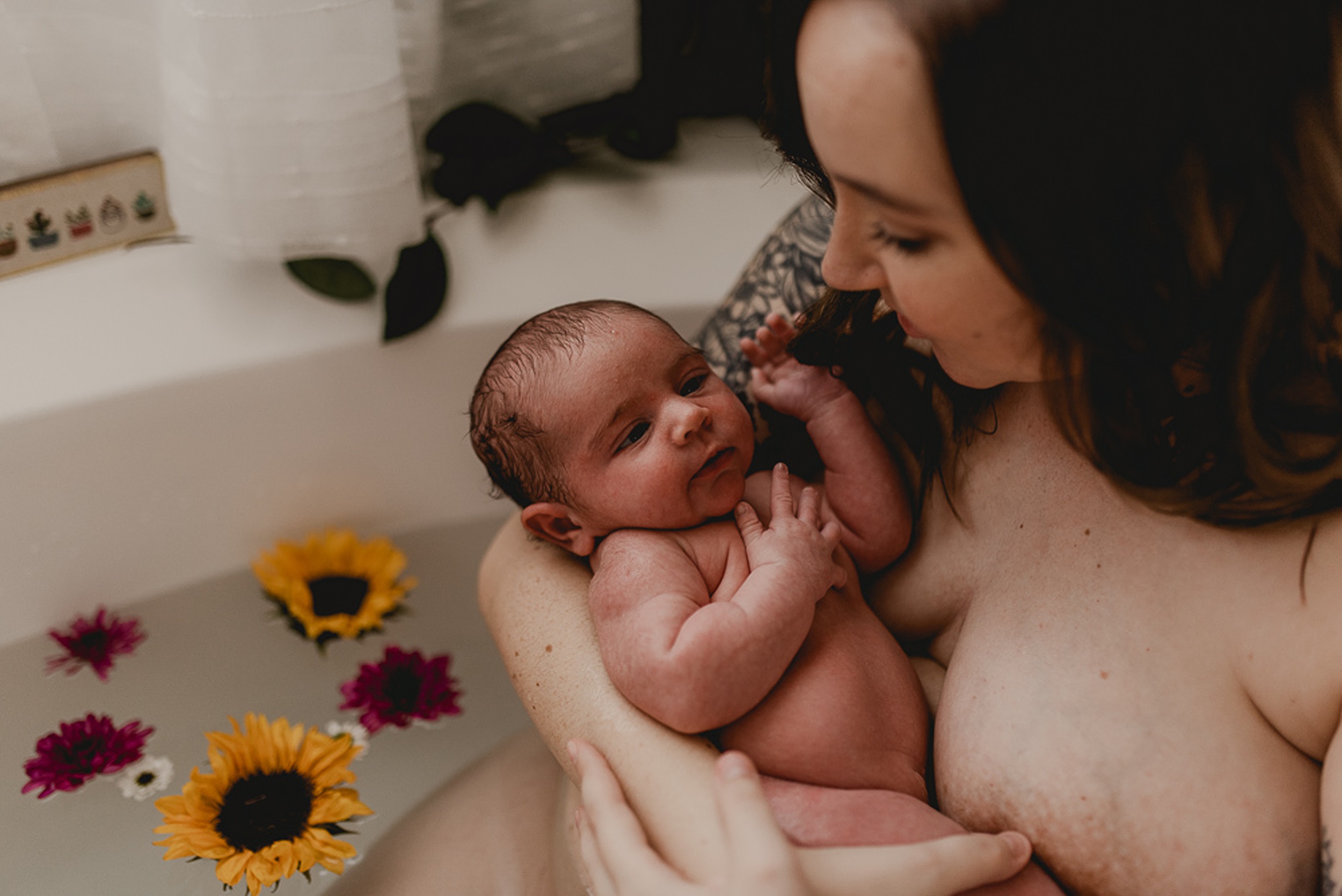 A mother smiles down at her newborn baby in her arms while sitting in a bathtub thanks to family first midwifery