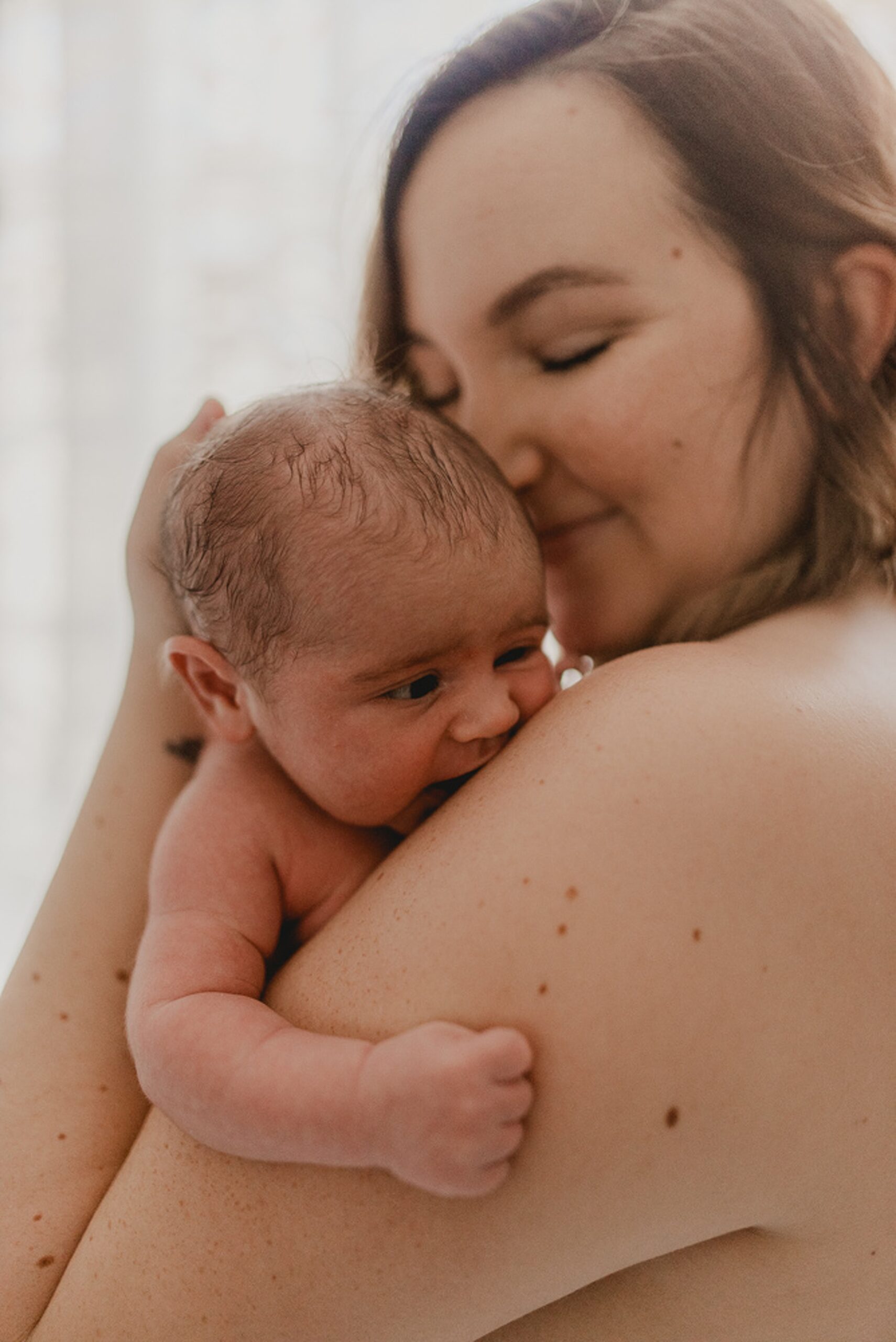 A mother stands in a bathroom cradling her newborn baby against her shoulder and smiling thanks to family first midwifery