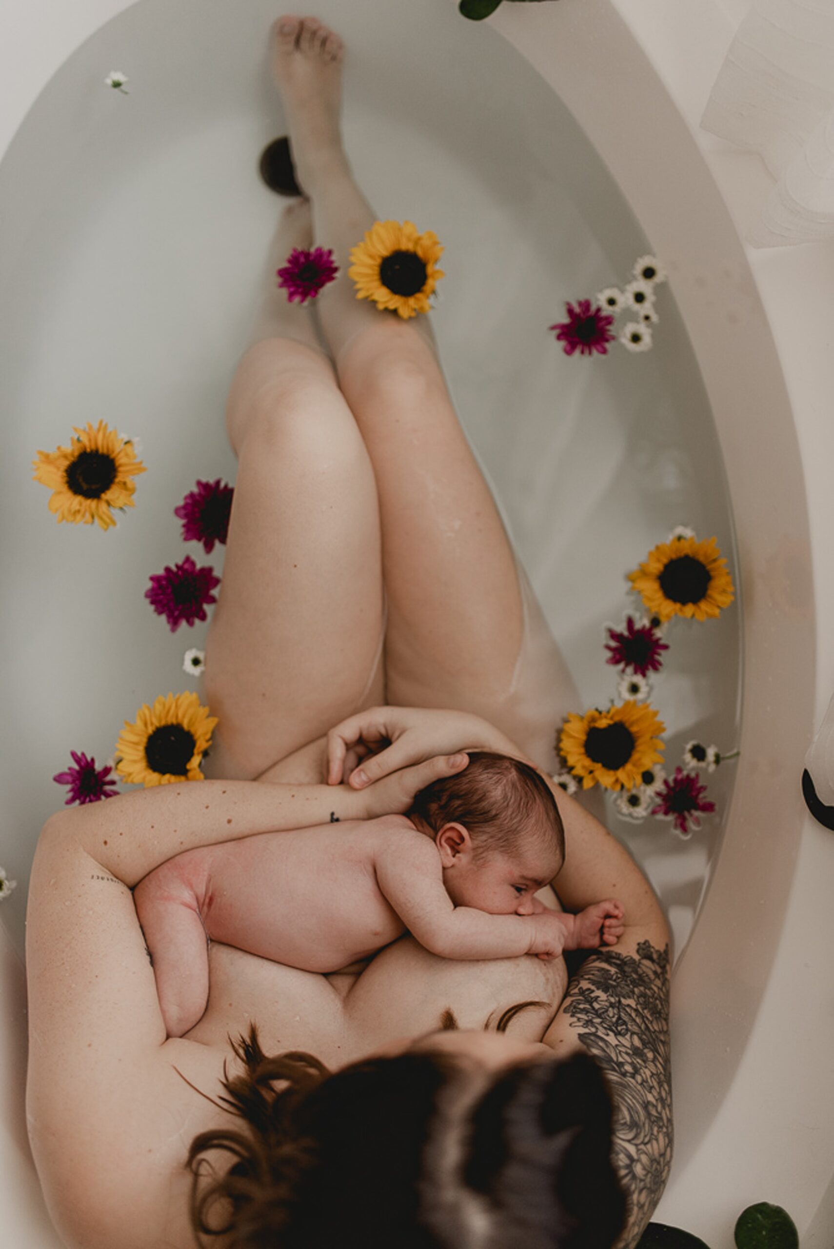 A new mom breastfeeds her newborn baby while sitting in a bathrub with flowers floating around them
