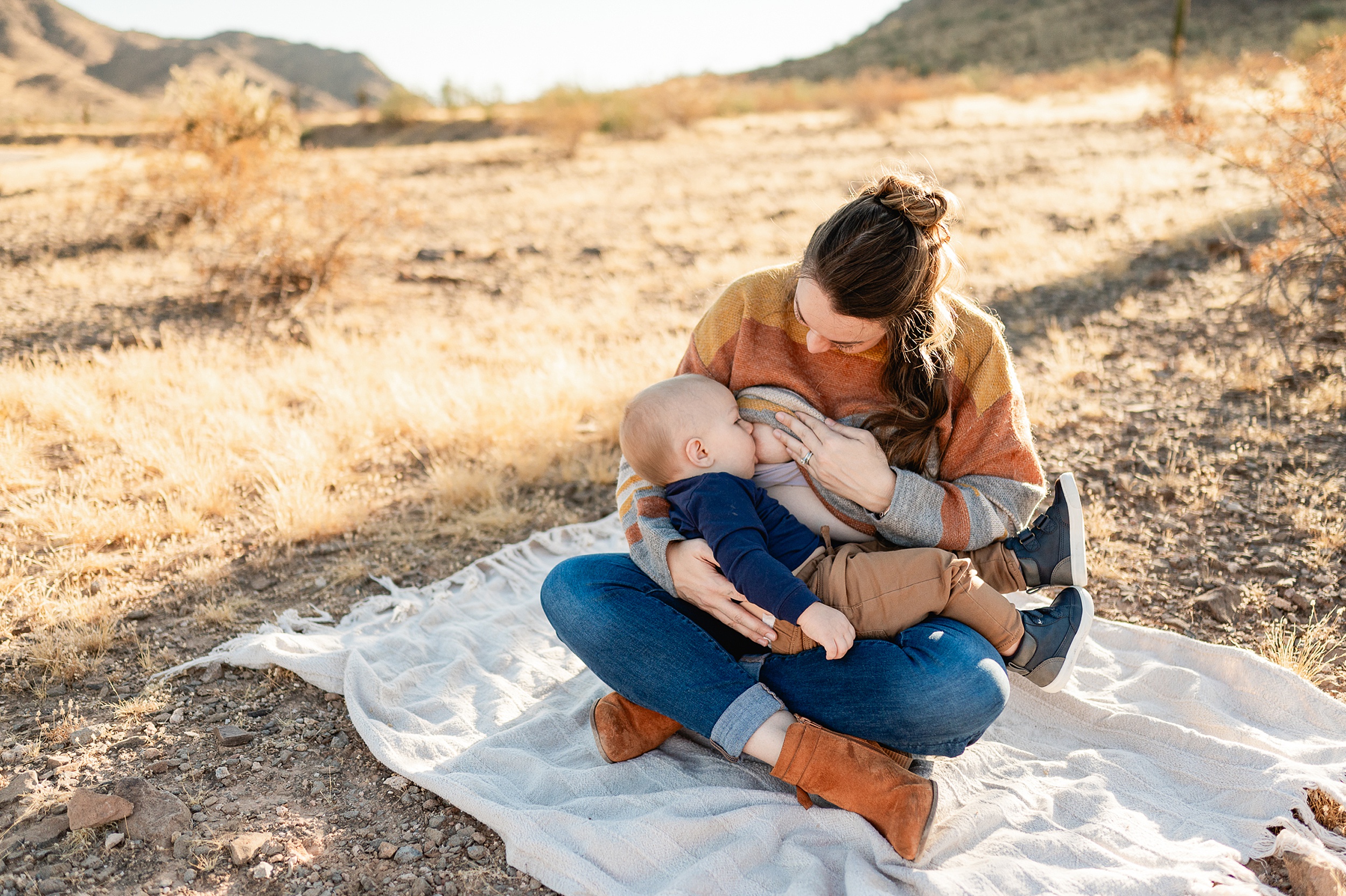A mother breastfeeds her toddler son while sitting on a picnic blanket in a desert park