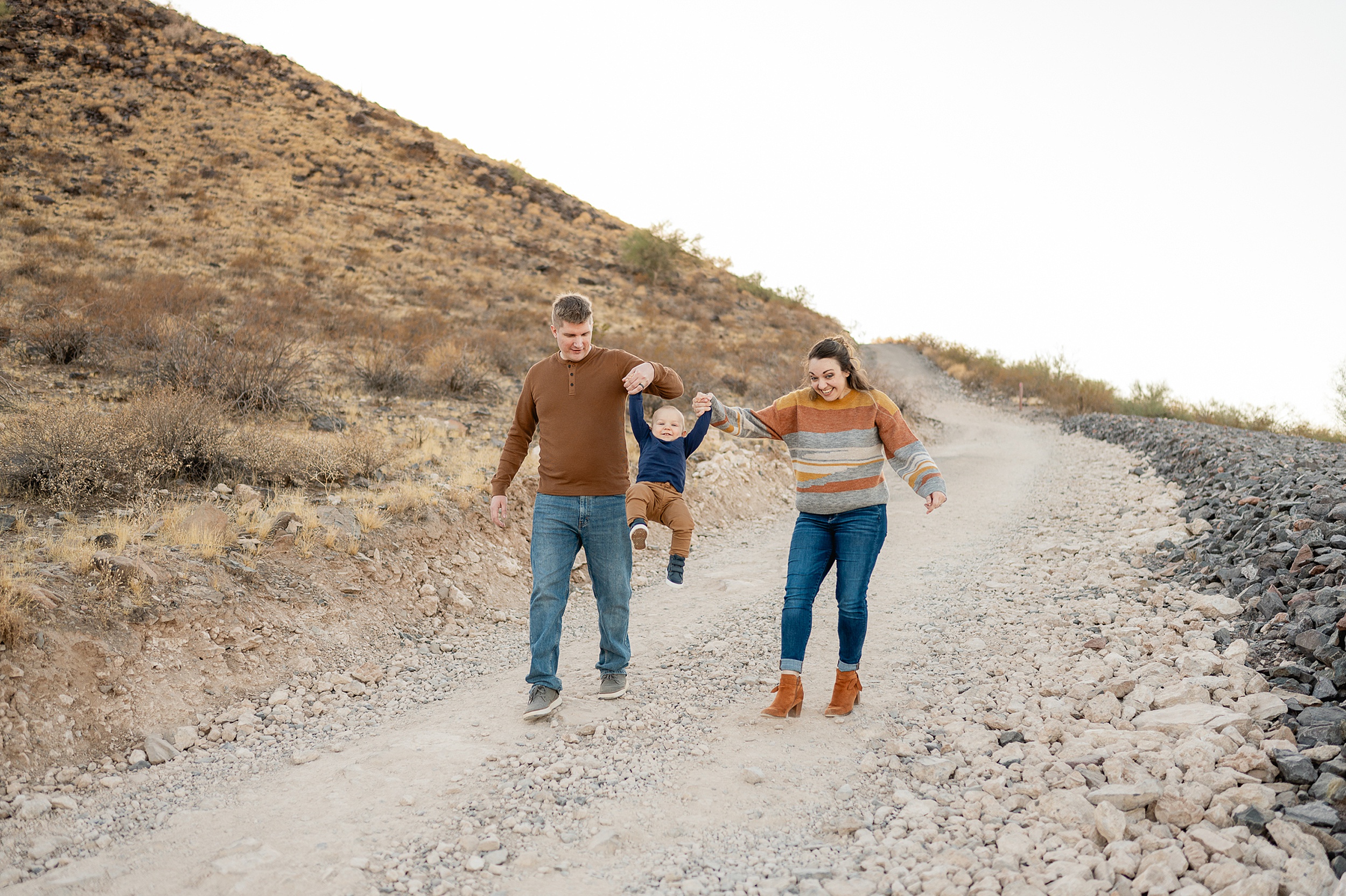 A mom and dad swing their toddler son while walking down a desert path