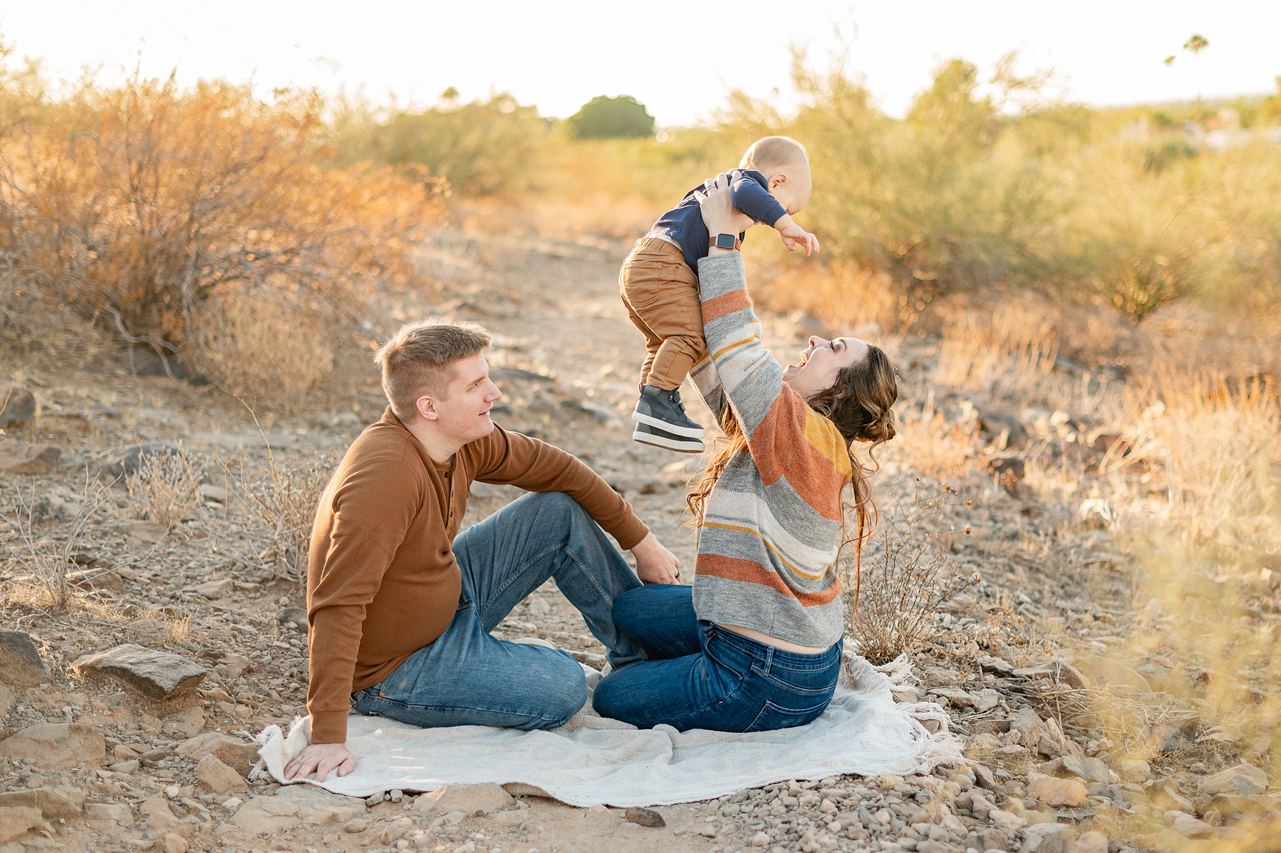 A mother and father sit on a picnic blanket in a desert trail at sunset playing and laughing with their toddler son before visiting scottsdale yoga studios