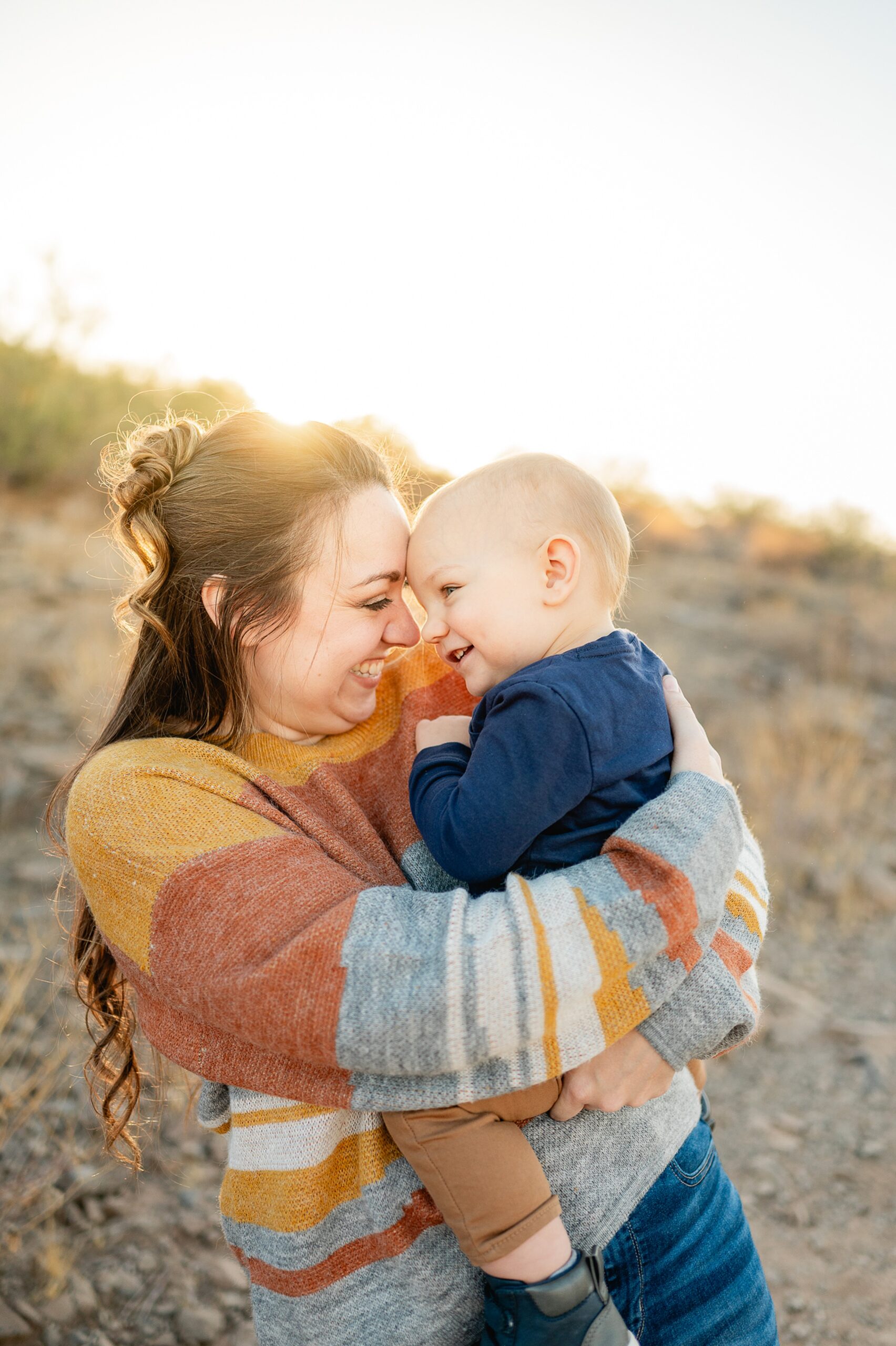 A happy mother in a colorful sweater laughs and touches foreheads with her toddler son on her hip at sunset in a park after visiting scottsdale yoga studios