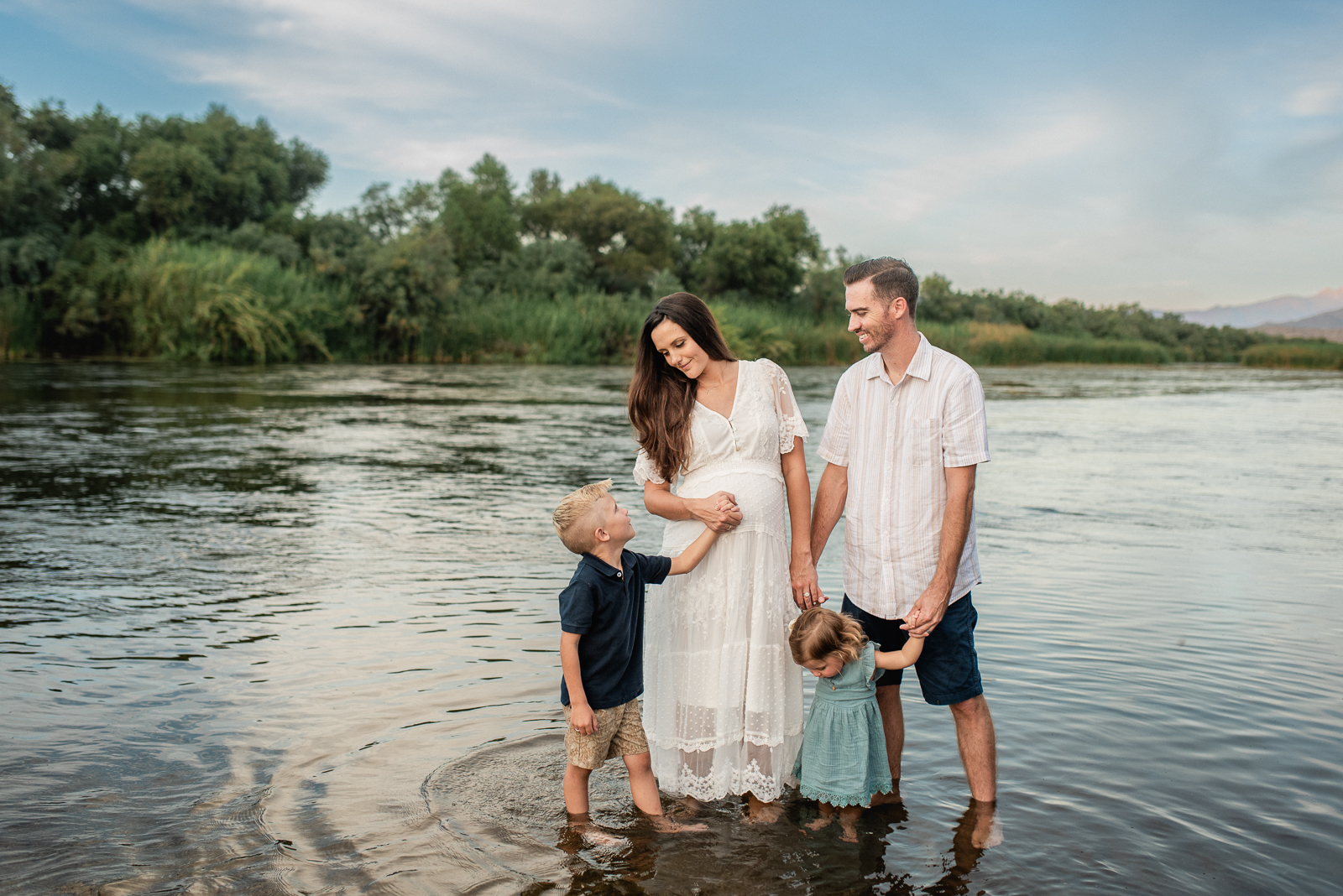 a family of four standing at the edge of the Salt River while pregnant mom in a beautiful white dress holds the hands of her two little kids and her husband looks on