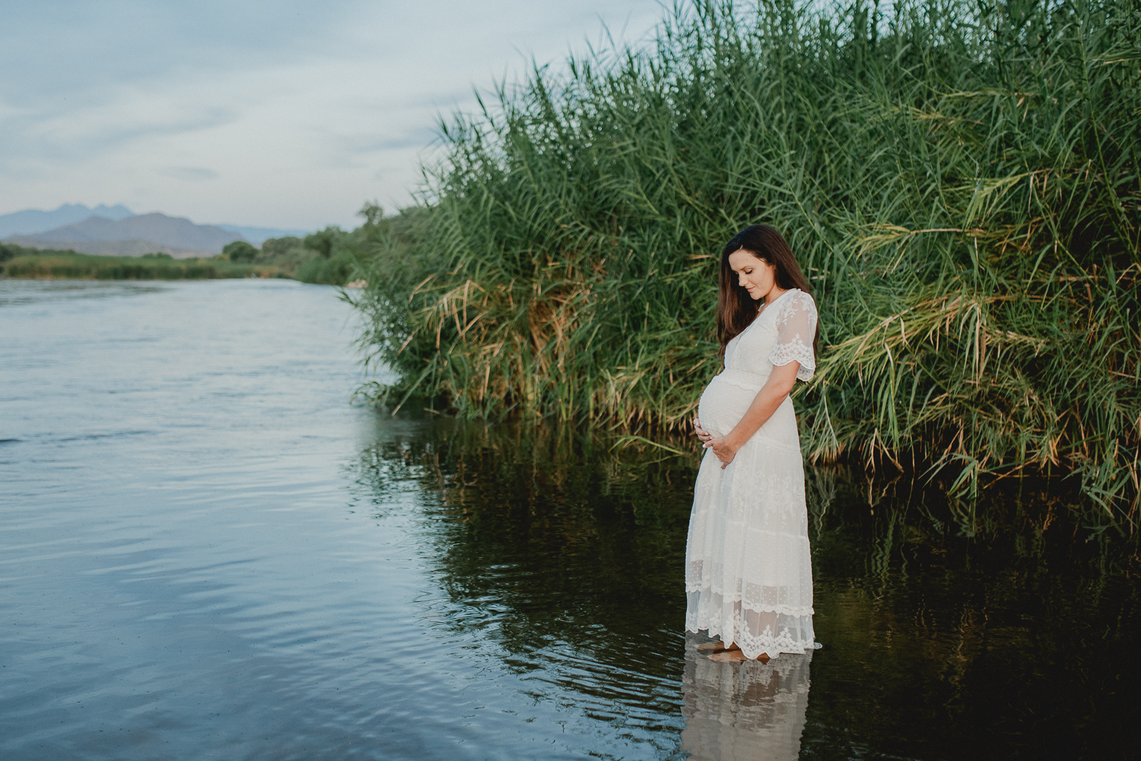 Pregnant mother stands in the Salt River holding her tummy thinking about her support team at cherry blossom doulas