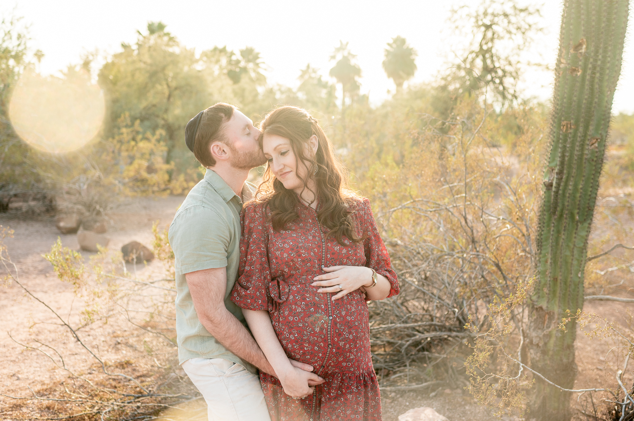 pregnant wife holding her tummy wearing a maroon floral dress while her husband holds her hand and kisses her temple at an outdoor maternity photo session