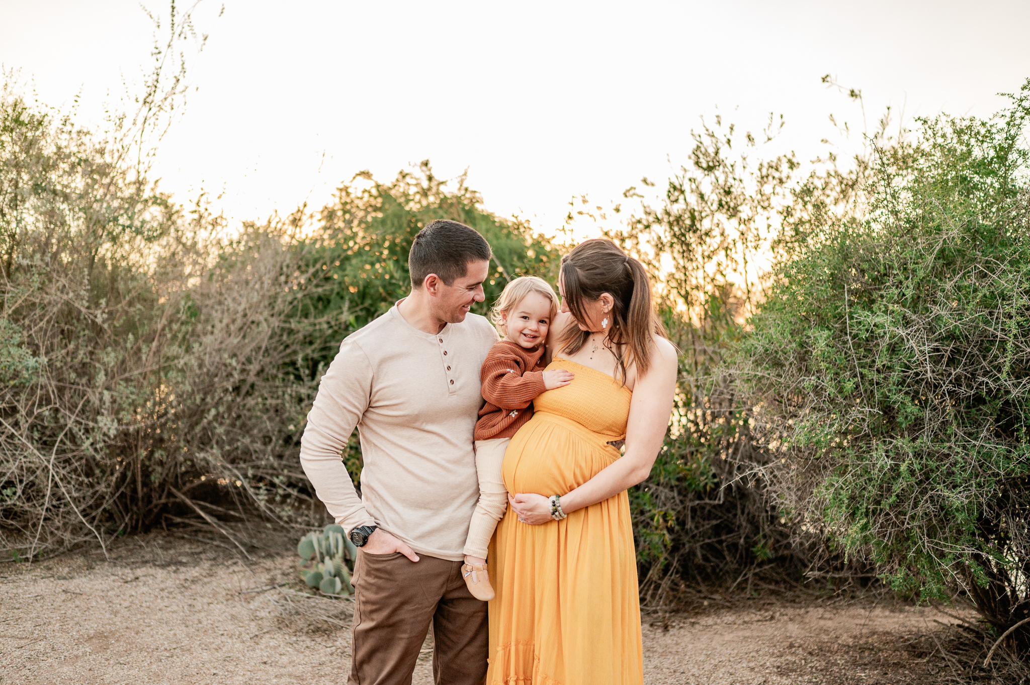 Family of three with pregnant mom in yellow gown holding her toddler girl in a rust sweater and dad in a cream shirt and brown pants holding the other two in front of Phoenix green backdrop