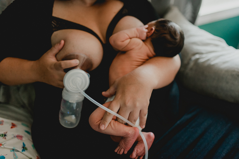 Mom holds newborn baby who is naked to one breast while he nurses and she pumps breastmilk on her other breast