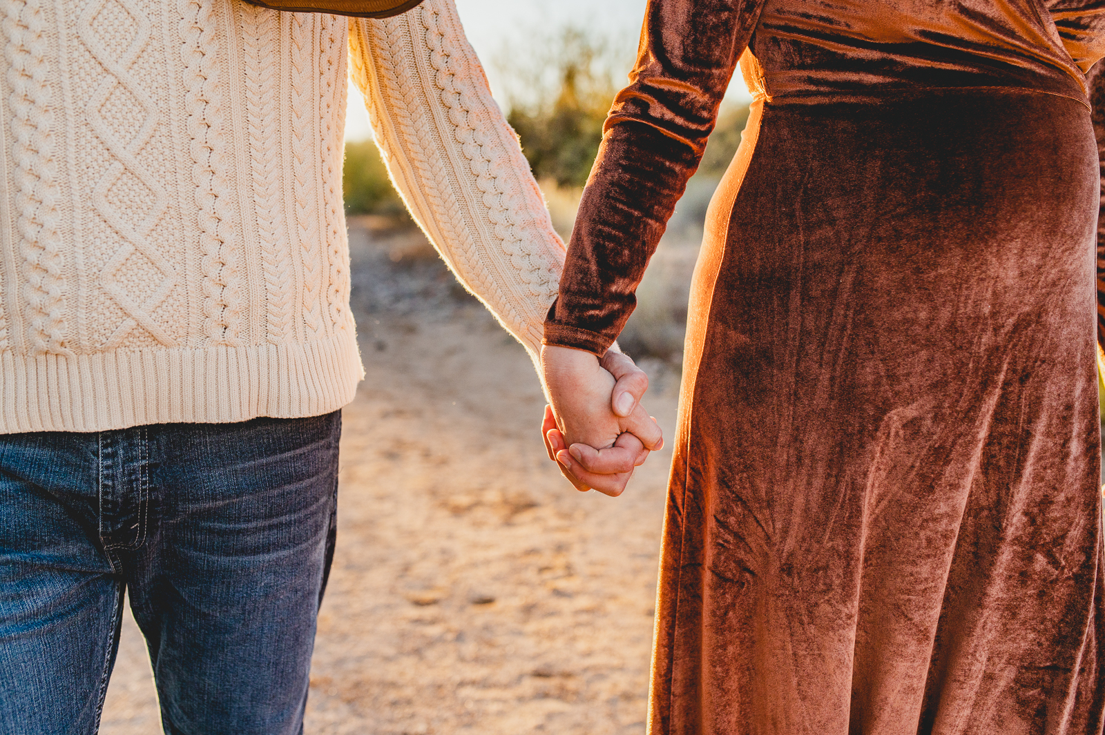 husband wearing dark jeans and a cream sweater holds hands with his pregnant wife who's wearing a velvet rust maxi dress at her desert maternity photoshoot