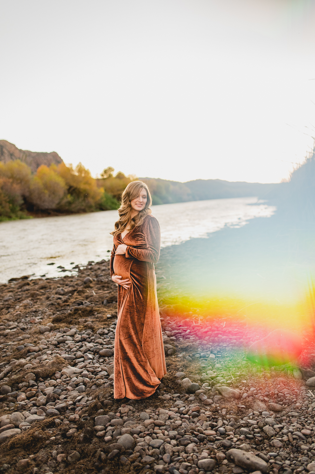 pregnant woman in a rust orange velour maternity dress stands on the rocks beside the Salt River in Phoenix looking off to her side while holding her pregnant belly amidst rainbow shafts of light