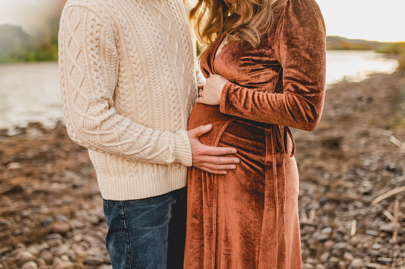 husband holds his wife's pregnant belly on the bottom while she holds it on top. he is wearing jeans a cream sweater and she wars a velour maxi dress in deep rust as they stand by the bank of a Phoenix river