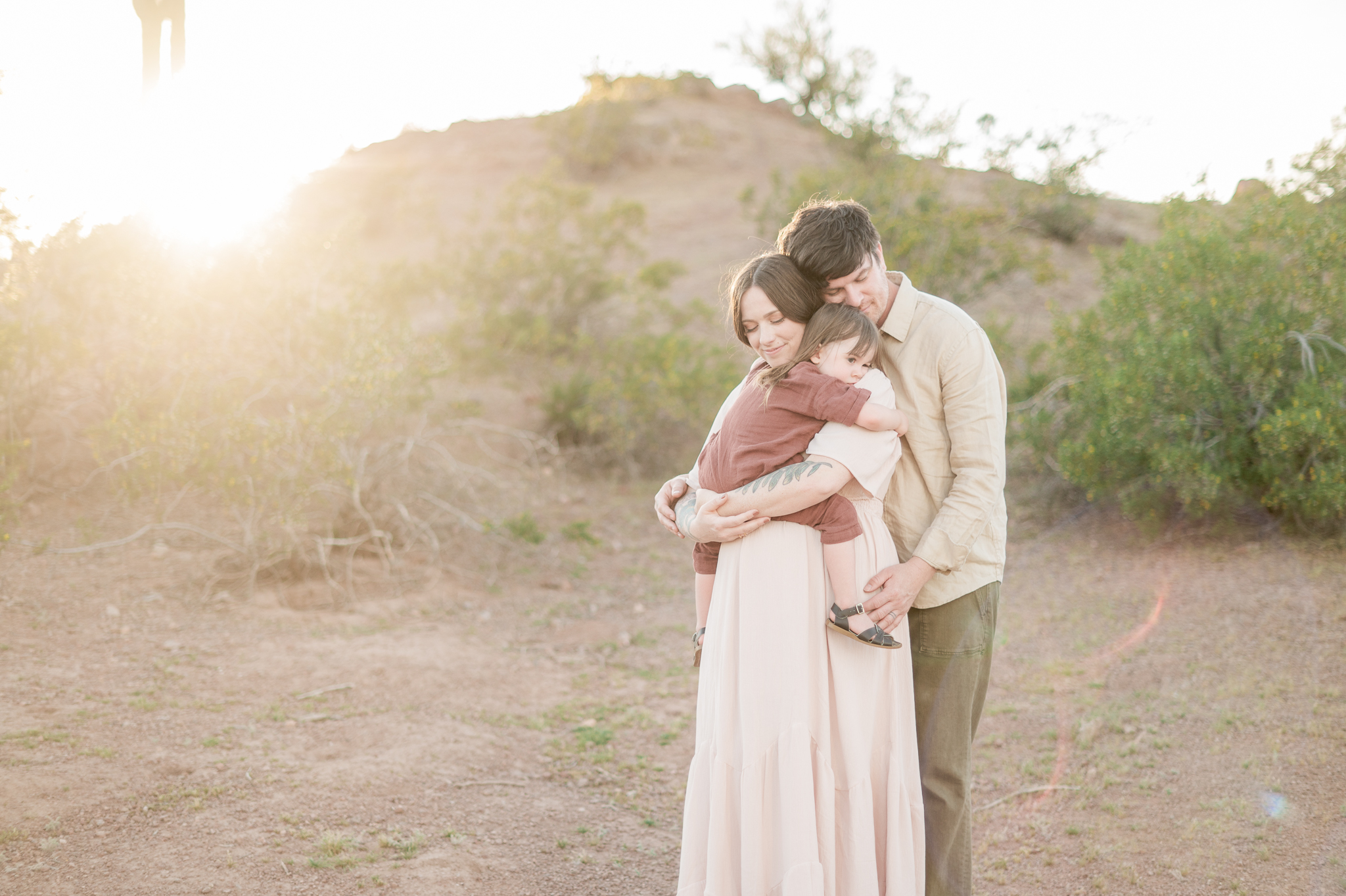 pregnant mama in a pink gown holds her toddler in a rust romper while dad holds on to both of them from behind with the golden sunlight shining behind them