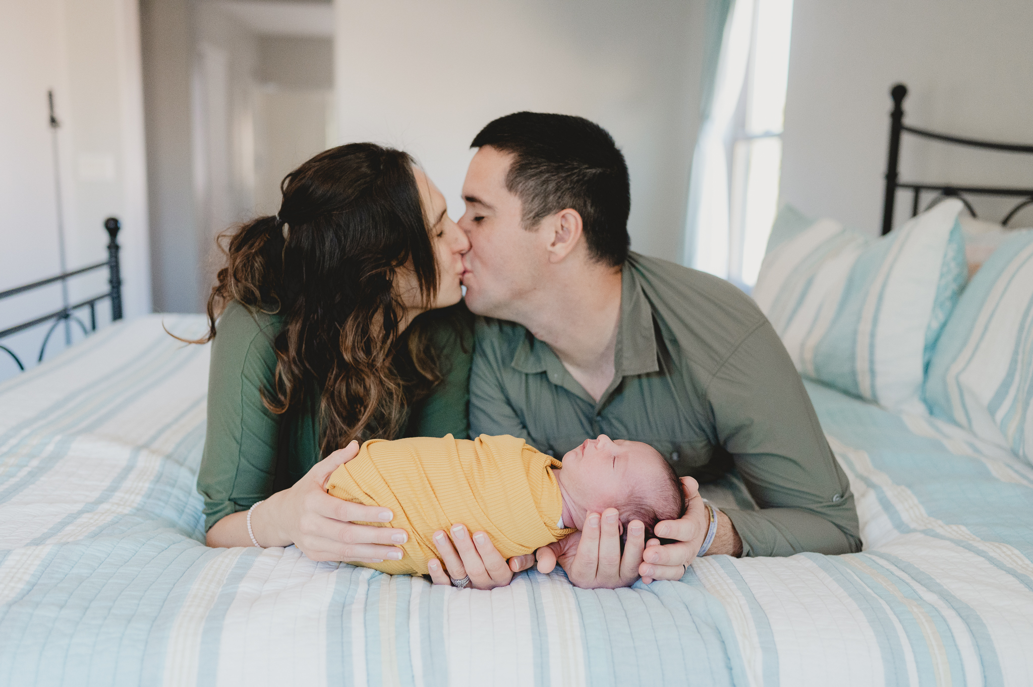 comfortable indoor newborn photography session where mom and dad are on their stomachs on the bed holding baby in front of them and kissing each other