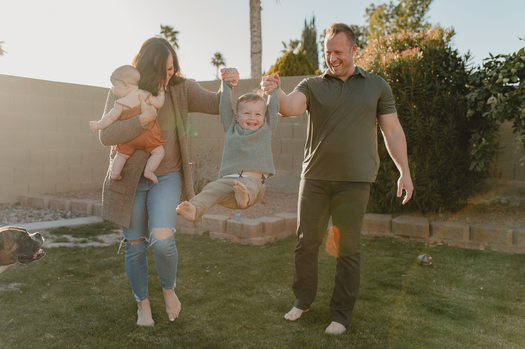 mom, holding baby girl, and dad both hold their toddler sun and swing him in their backyard in home session in Scottsdale, AZ 