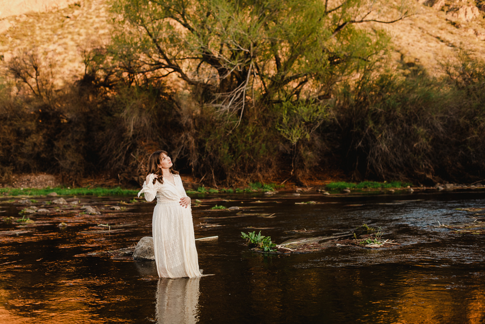 A pregnant woman in a white dress walks through a flowing shallow river at sunset in the desert