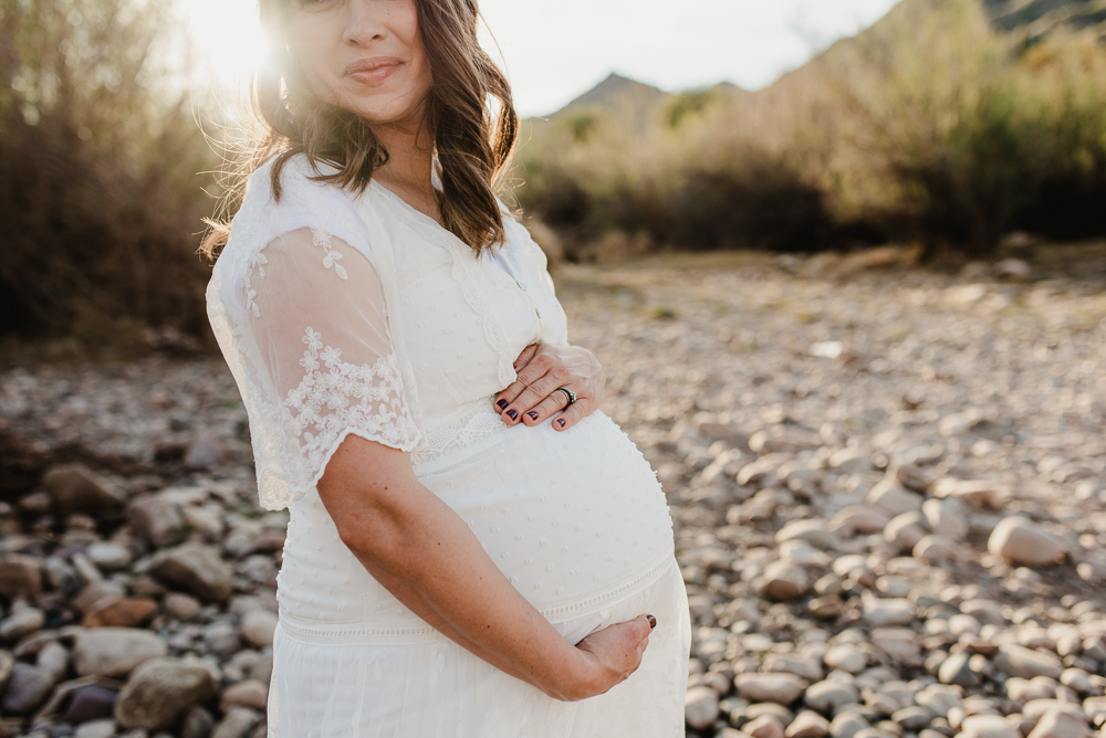 A mother to be in a white dress smiles while holding her bump in a dry riverbed at sunset after meeting phoenix doulas