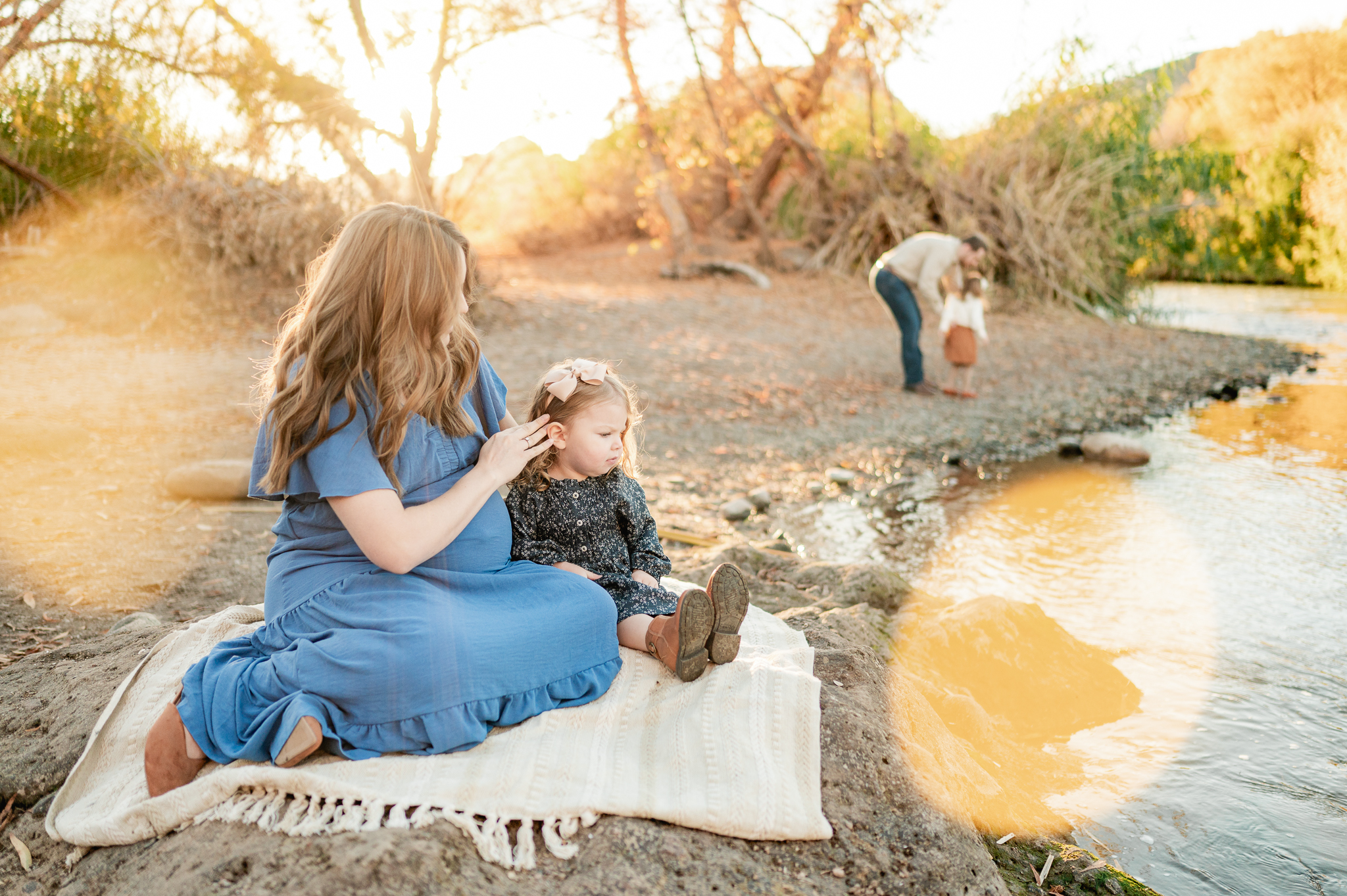 mother sits on a blanket with her young daughter at Phon D Sutton as dad dances with their older daughter in the background at the bank of the river