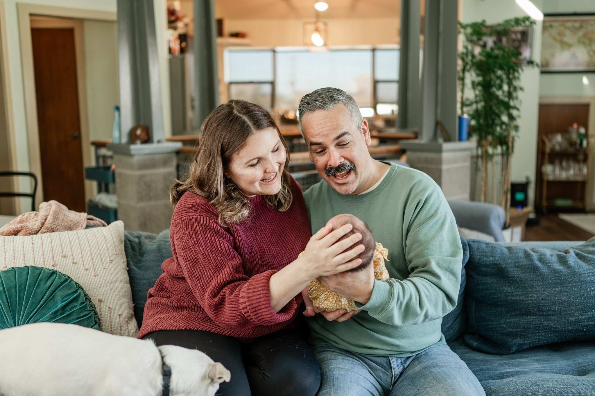 Happy new parents sit on a couch smiling with their newborn baby in dad's hands between them after visiting strolleria scottsdale