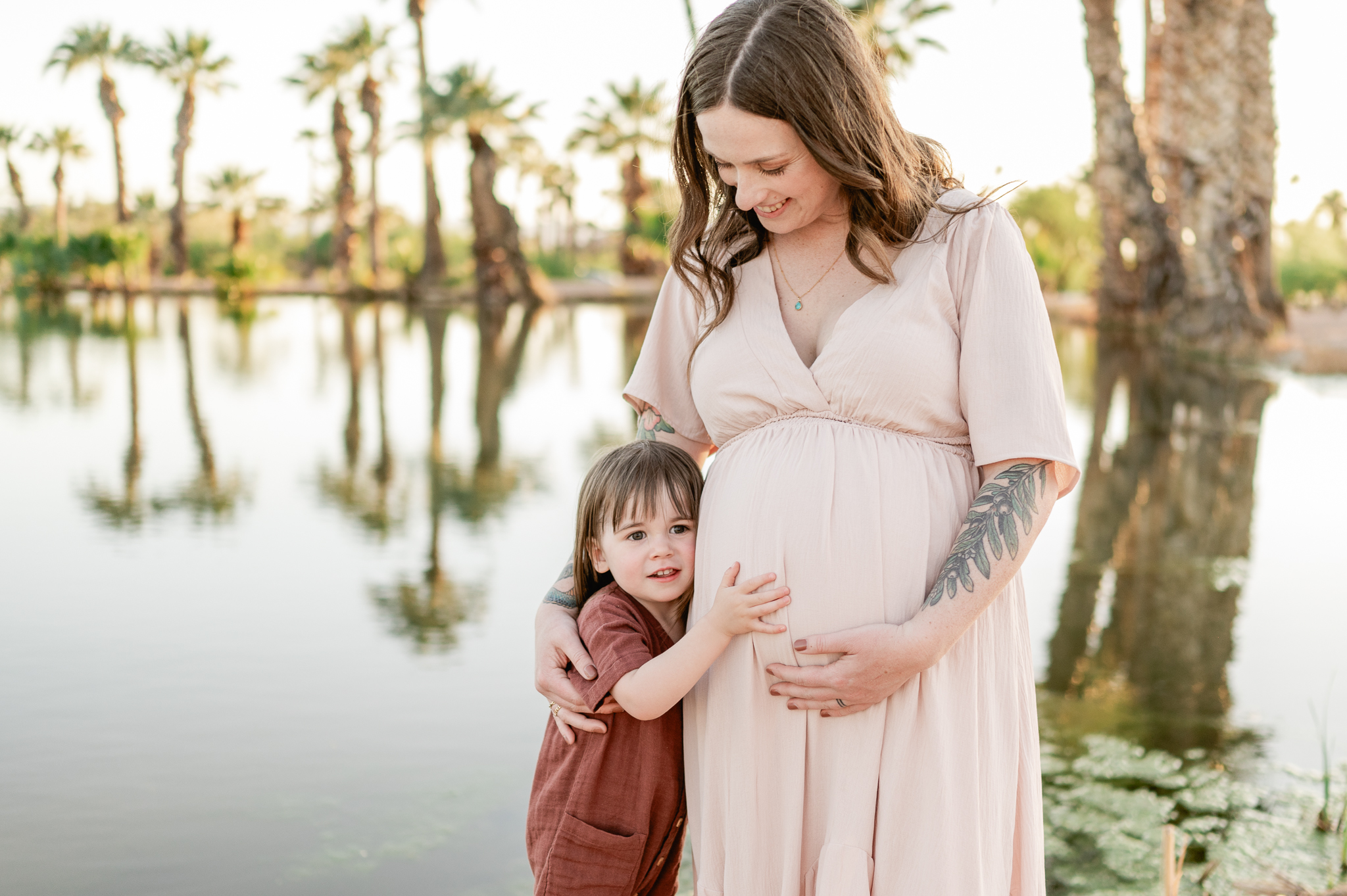 pregnant mom in a light pink dress holding her tummy while bringing her toddler girl close to her in an embrace