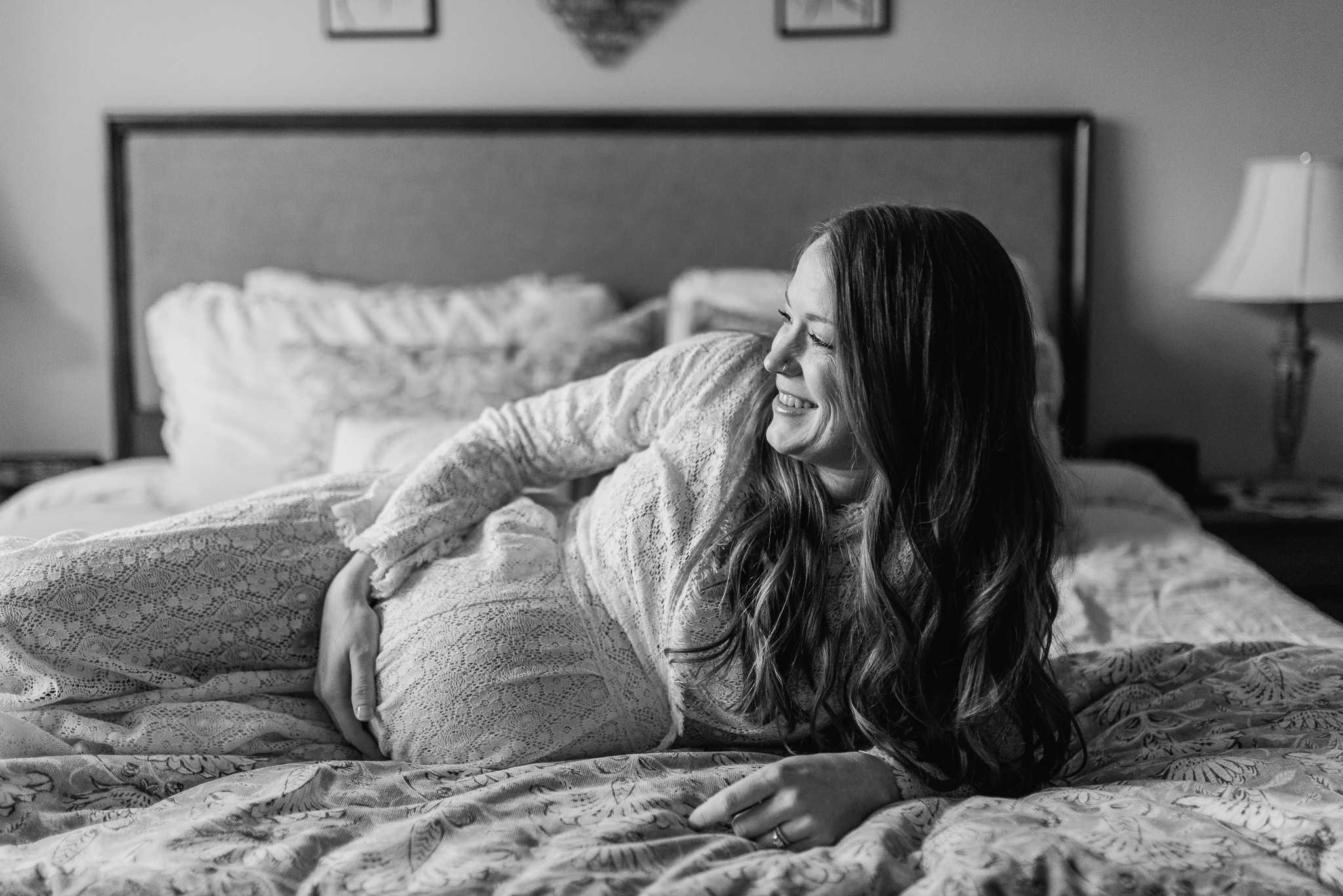 black and white image of pregnant mom lying on her side on her bed looking out the window smiling