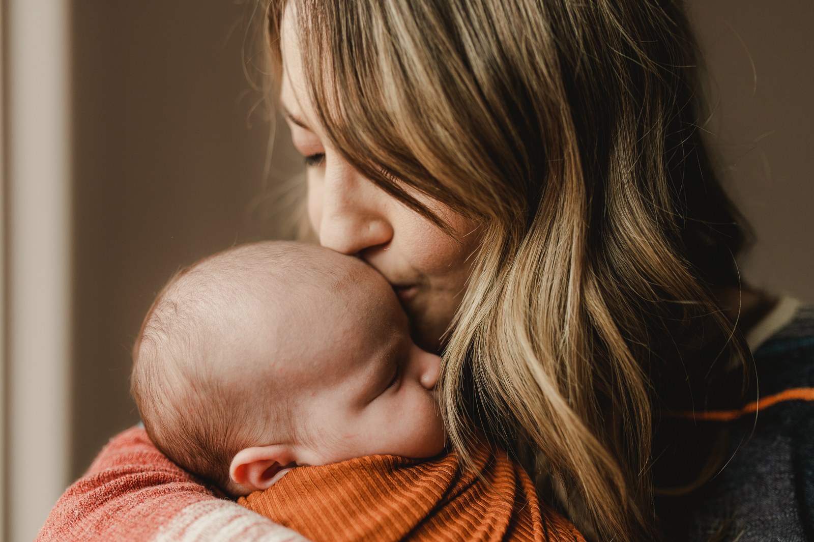 mom kissing the head of her newborn baby girl swaddled in a rust cotton wrap